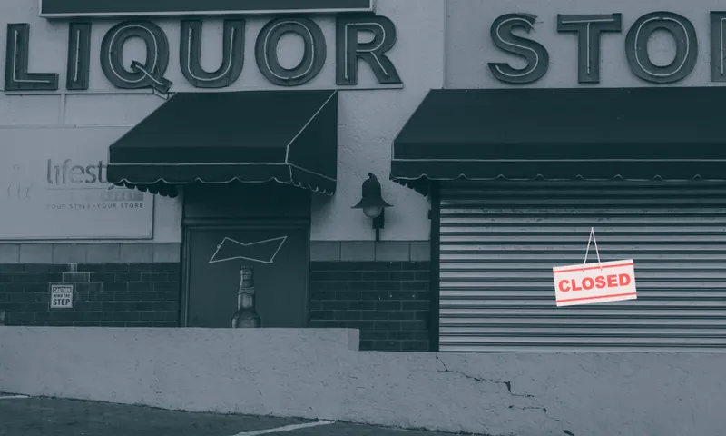 A liquor store with closed metal shutters and a "CLOSED" sign hanging on the front. The entrance and windows are covered, and the building exterior has large store signage.