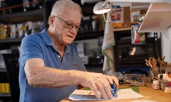 A man in a blue polo shirt sits at a cluttered desk, concentrating as he works on a small craft project under a lamp in a cozy, well-lit workspace.