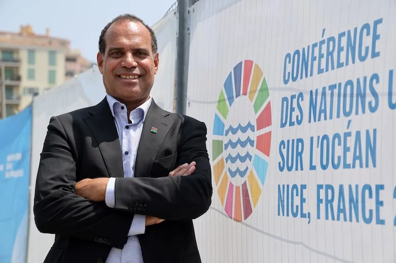 A man in a black suit stands smiling with arms crossed in front of a fence displaying the United Nations Ocean Conference logo and text reading “Conférence des Nations Unies sur l’Océan, Nice, France.”.