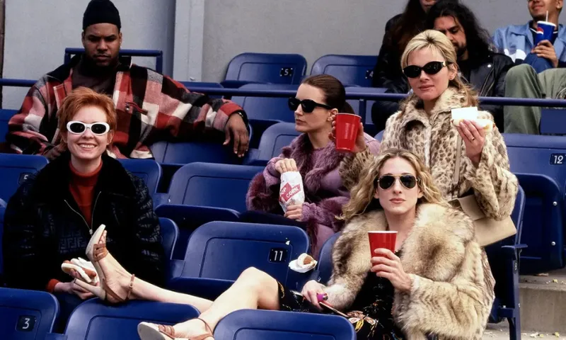 Four stylish women sit in stadium seats, wearing sunglasses and fashionable coats, holding drinks and snacks, with empty blue seats around them and other people in the background.