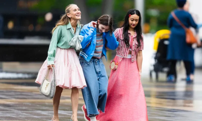 Three women walk together outdoors, smiling and laughing. They wear colorful, stylish outfits, including skirts and jeans, and appear to be enjoying each other's company on a city street.