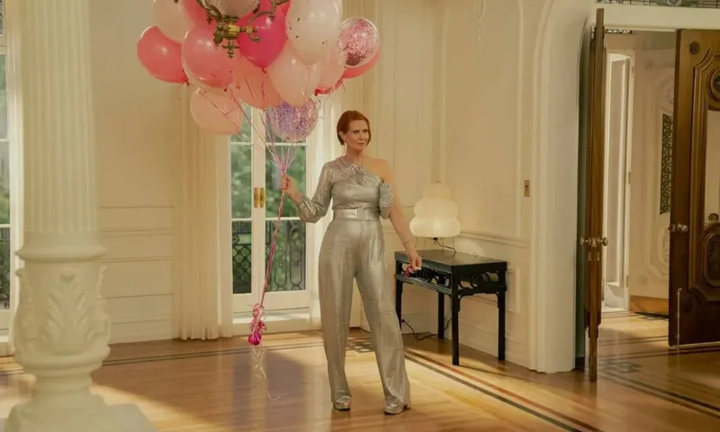 A woman in a shiny silver jumpsuit stands in a bright, elegant room holding a large bunch of pink and white balloons. Sunlight streams through tall windows, and the room features classic white decor and wooden floors.