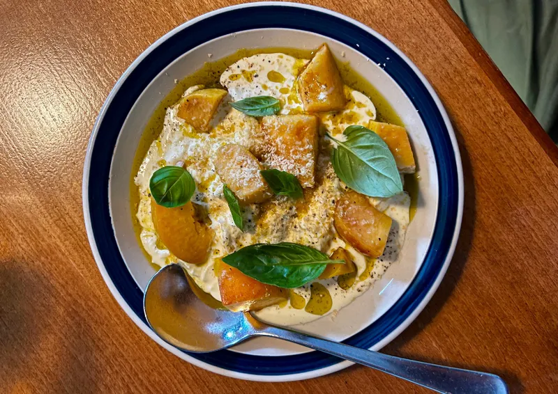 A bowl of creamy soup topped with golden roasted root vegetables, fresh basil leaves, grated cheese, and a drizzle of oil sits on a wooden table with a metal spoon resting on the side of the dish.