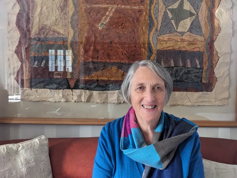 Kate Dewes, an older woman with grey hair and a colourful scarf, sits on a red/brown sofa with some earth-toned art behind her and a slight smile