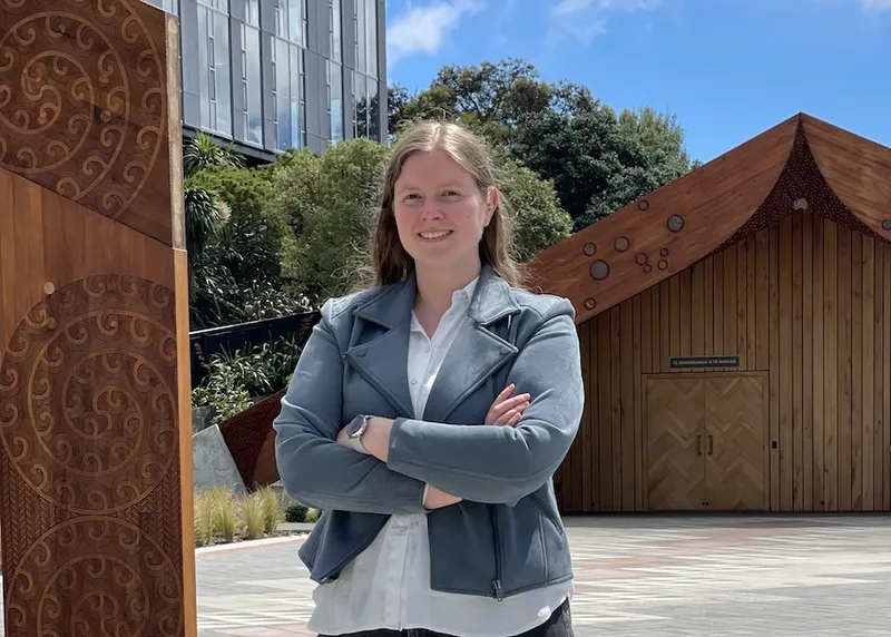 Lauren Vinnell, a woman wearing a blue jacket, stands outside with her arms crossed in the sun. she has light skin and hair
