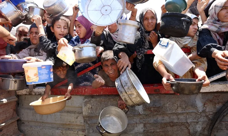 A crowd of people, including children and adults, reach out with empty pots and containers, appearing eager and desperate, as they wait behind a barrier, suggesting a scene of food distribution or aid relief.