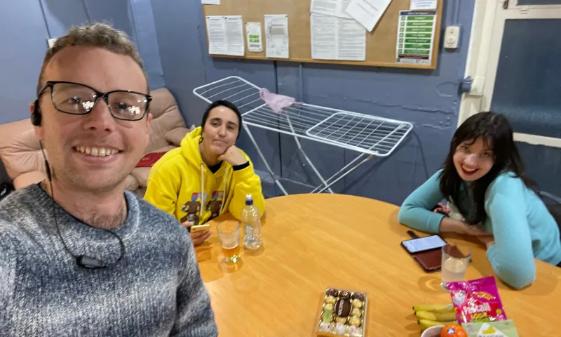 Three people sit around a wooden table with snacks, drinks, and a phone. One person takes a selfie, smiling, while the other two also smile at the camera. An empty drying rack and bulletin board are in the background.