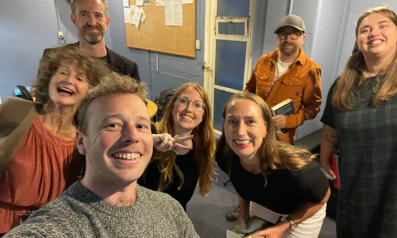 A group of seven smiling adults pose for a selfie in an indoor room with blue walls and a bulletin board in the background. Some people hold books, and one person flashes a peace sign.