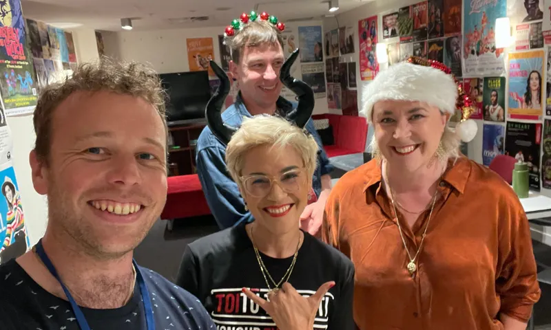 Four people smiling indoors, some wearing festive accessories: one has reindeer antlers, another black horns, and another a Santa hat. They stand in front of a wall covered with colorful posters.