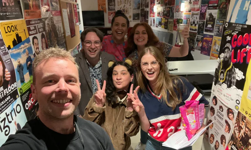 A group of six people smile and pose for a selfie in a hallway covered with colorful event posters. One person is holding up a peace sign, and another is holding a snack bag and a drink. The atmosphere is cheerful and lively.