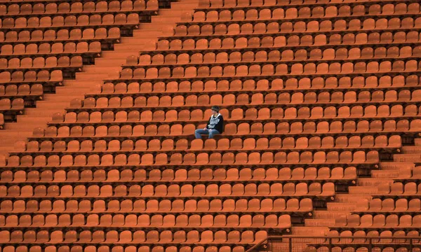 A lone person sits among many empty orange seats in a large stadium, appearing small and isolated in the vast, deserted space.
