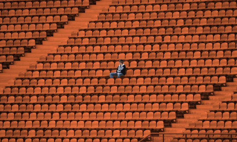 A lone person sits among many empty orange seats in a large stadium, appearing small and isolated in the vast, deserted space.