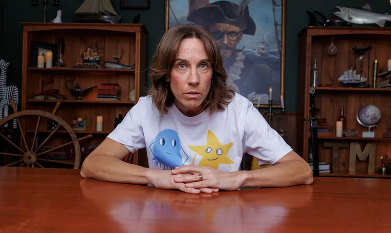 Jackie wears a white tshirt and stares down the camera in front of assorted naval objects