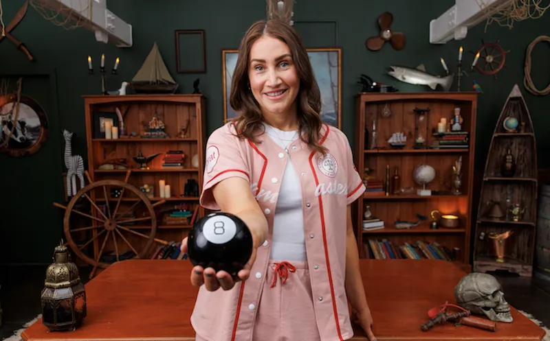 Bree wears a baseball outfit with long brown hair and holds a number eight ball towards the camera.