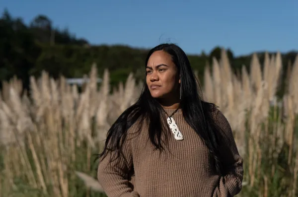 A woman with long dark hair wearing a brown sweater and a carved necklace stands outdoors in front of tall grasses, looking thoughtfully into the distance under a clear blue sky.