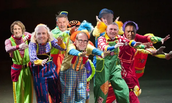 A group of people in colorful clown costumes pose together on stage, each with a unique outfit, bright wigs, and exaggerated makeup, striking playful and animated poses.