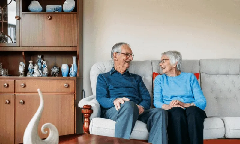 An elderly man and woman sit together on a light-colored sofa, smiling and talking. Behind them is a wooden cabinet with decorative items. The room looks warm and inviting.