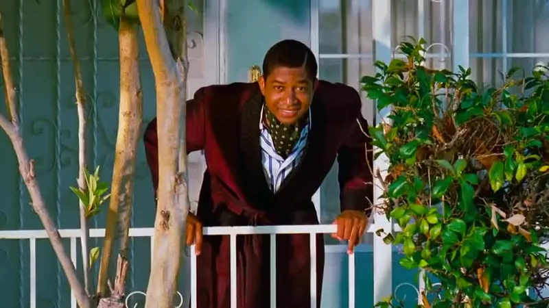 A man wearing a dark robe and patterned scarf leans over a white railing on a porch, surrounded by plants and greenery, with a house and window in the background.