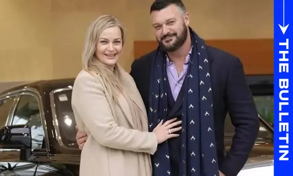 A smiling couple stands in front of a car, with the woman wearing a beige coat and the man in a dark suit and patterned scarf; "THE BULLETIN" is written vertically on a blue banner on the right side.