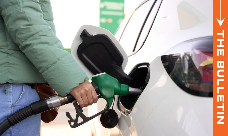 A person wearing a green jacket is refueling a white car at a gas station. The fuel nozzle is green. On the right side, an orange vertical banner reads "THE BULLETIN.