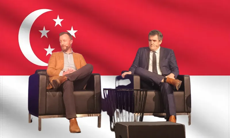 Two men in suits sit on black armchairs on a stage with the Singapore flag in the background. A small black table is between them, and a black box is in front of the stage.