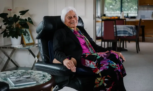 a woman with brown skin and white hair sits in an armchair at home, smiling at the camera