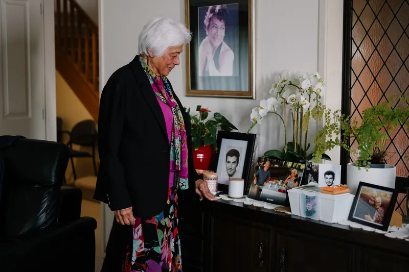 an older woman with white hair stands next to a side table looking at framed photos. she wears a colourful long dress and a black blazer