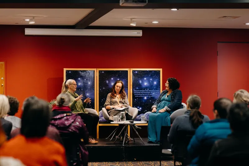 Three people sit on a stage in front of an audience, engaged in a panel discussion. Behind them are posters with starry night images. The stage and walls are red, and the room is warmly lit.