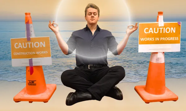 A man sits cross-legged and meditates on a beach at sunset between two orange caution cones with signs reading "CAUTION CONSTRUCTION WORKS" and "CAUTION WORKS IN PROGRESS." A glowing halo circles his head.