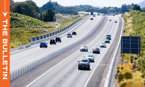 Several cars travel in both directions on a wide, divided highway surrounded by greenery and trees, with a large blank road sign and an orange sidebar reading “THE BULLETIN” on the left.