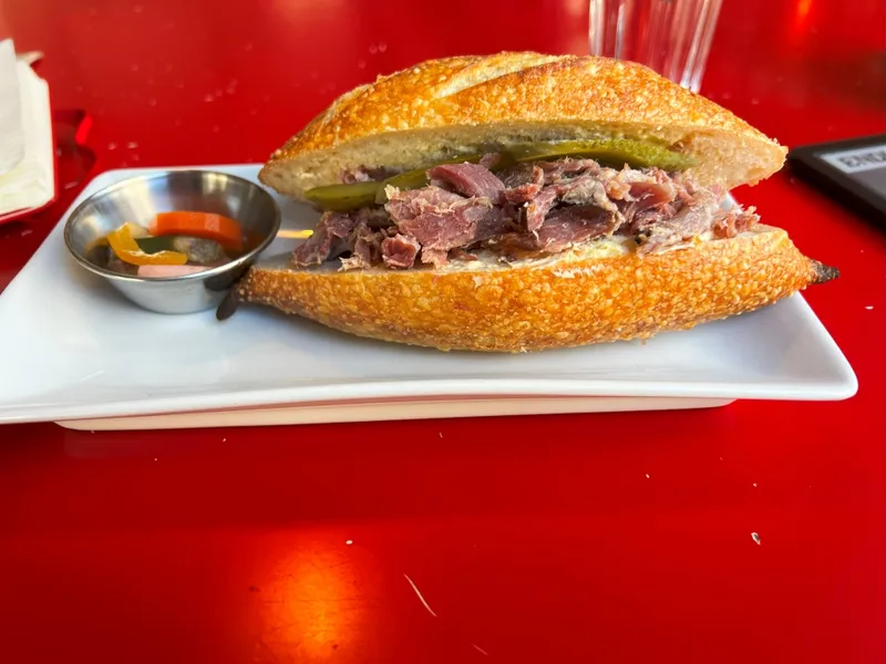 A sandwich with shredded meat and pickles on crusty bread sits on a white plate, next to a small dish of mixed pickled vegetables, on a bright red table. A glass of water and napkins are in the background.