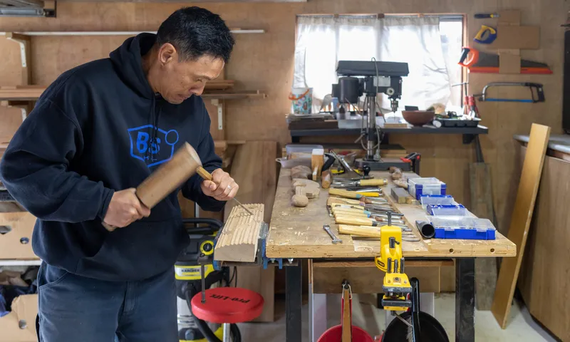 A man in a workshop uses a wooden mallet and chisel on a piece of wood at a workbench covered with tools, with woodworking equipment and supplies visible in the background.