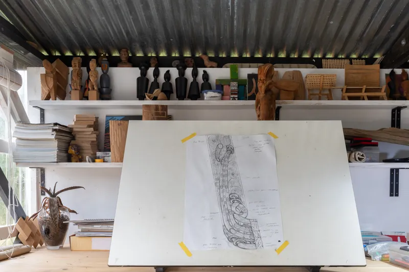 A sketch of a whakairo design is pinned to a tilted drawing board inside Leon Kipa’s studio. Shelves above hold carved figurines, timber samples, and small sculptural works in progress.