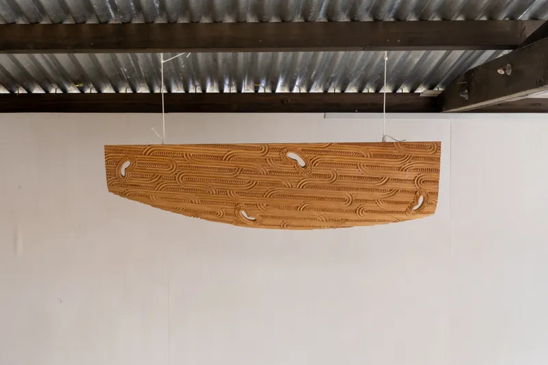 A large, cloud-shaped wooden sculpture — part of Leon Kipa’s Kapua series — hangs from the ceiling of his studio. Carved patterns run across its length, suspended against a plain white wall.