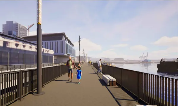 People walk along a wide waterfront promenade with railings, modern buildings to the left, and cranes and ships visible across the water under a partly cloudy sky.