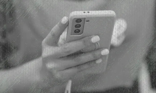 A person with manicured nails holds a smartphone with three rear cameras, using both hands. The image is in black and white with a textured, dotted overlay effect.