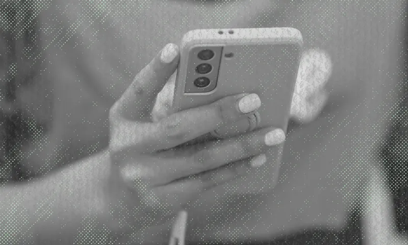A person with manicured nails holds a smartphone with three rear cameras, using both hands. The image is in black and white with a textured, dotted overlay effect.