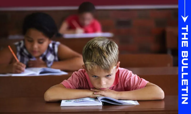 A young boy sits at a classroom desk, looking frustrated with his chin resting on a book. Two other children in the background are writing in their notebooks. A blue sidebar reads "THE BULLETIN.