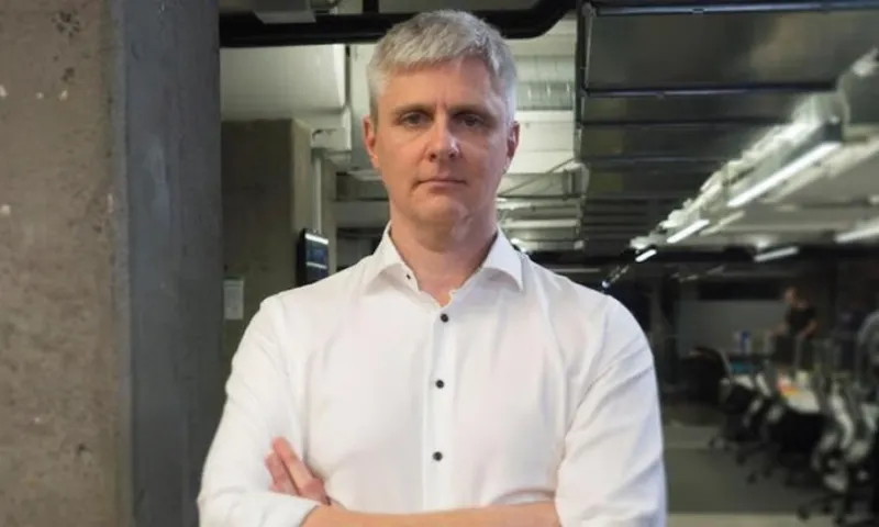 A man with short gray hair wearing a white button-up shirt stands with arms crossed in a modern office with industrial-style lighting and exposed concrete walls.