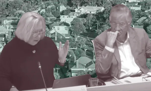 Two people sit at a desk in discussion, with one gesturing and the other resting their chin on their hand; a background of suburban houses is seen behind them.
