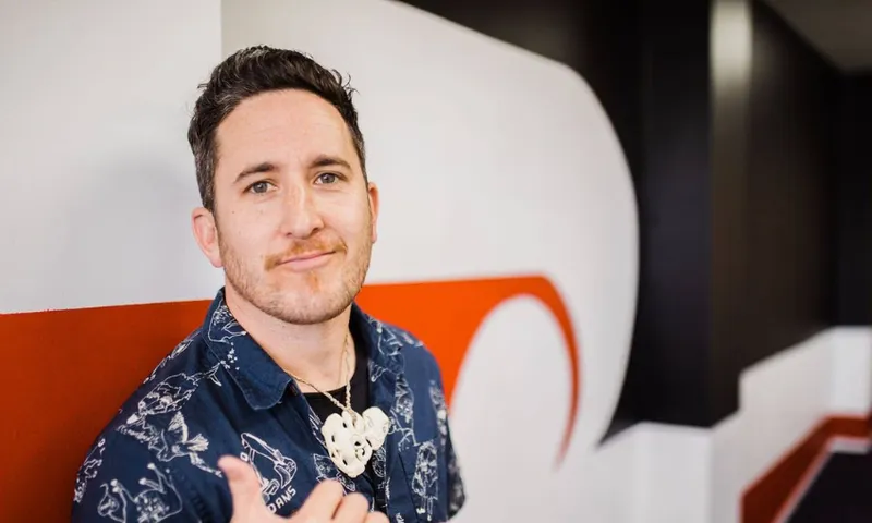 A man with short dark hair and a trimmed beard stands indoors, wearing a dark patterned shirt and a carved pendant necklace. He is smiling slightly and giving a shaka hand gesture, with a modern red, white, and black wall behind him.