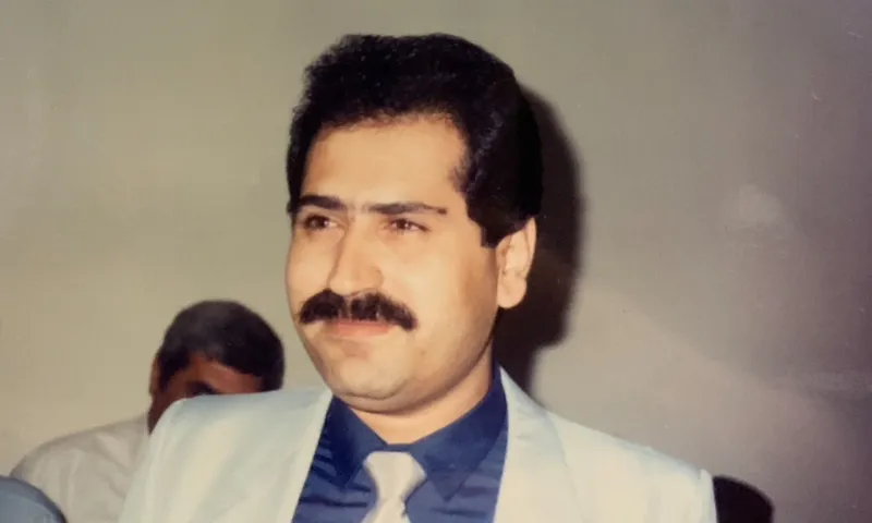 a man with dark hair and a dark moustache smiles off camera wearing a beige suit and blue shirt