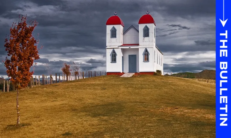 A small white church with two red domes stands on a grassy hill under a cloudy sky, with a single autumn tree nearby and a blue vertical banner on the right labeled "THE BULLETIN.