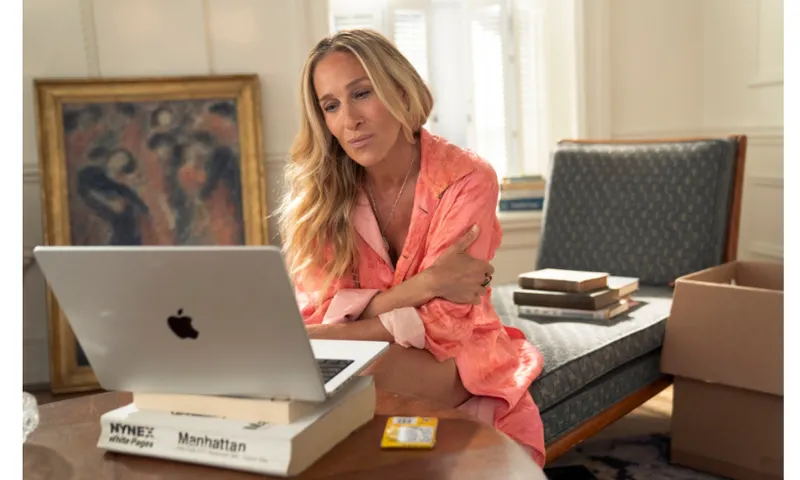A still from a TV episode showing Carrie Bradshaw at a her laptop which is on top of a large book. There's a painting and chaise lounge behind her as well as another stack of books. She is wearing a peach shirt.