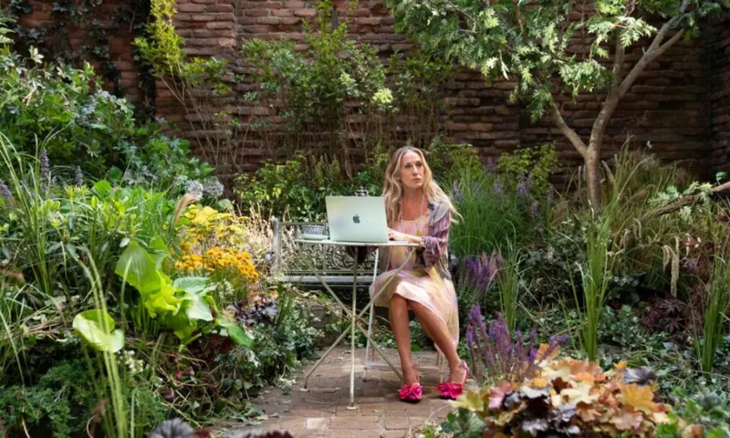 A still from a TV episode showing Carrie Bradshaw sitting at a table with her laptop outside in a lush garden.