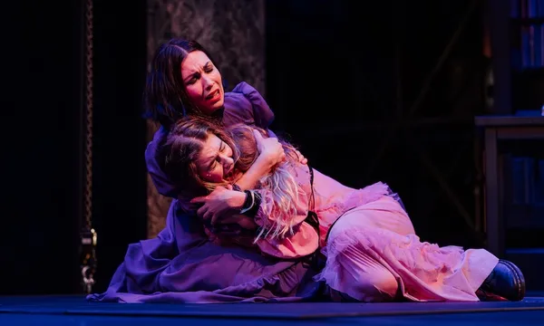 a woman with dark hair and wearing a purple dress holds a distraught woman in a pink dress. they are onstage