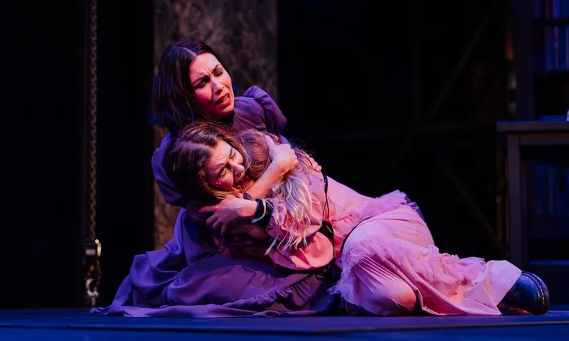 a woman with dark hair and wearing a purple dress holds a distraught woman in a pink dress. they are onstage