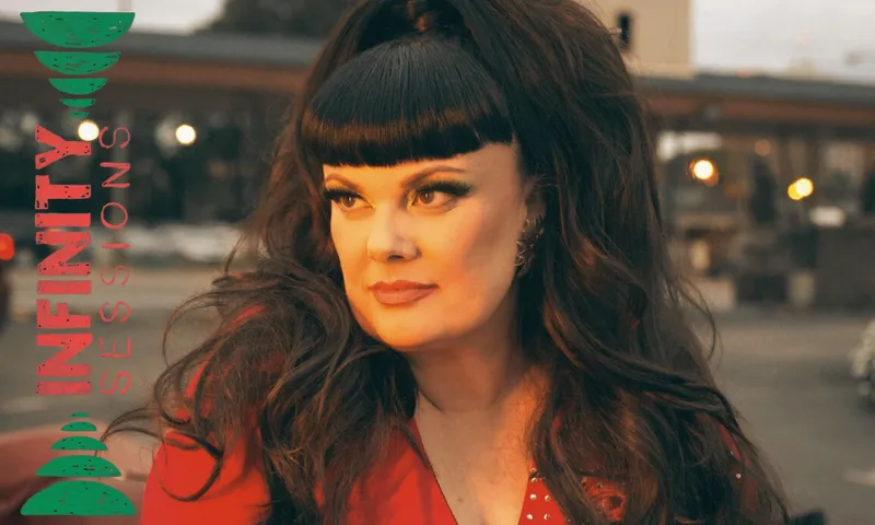 A woman with long dark hair and bangs, wearing a red outfit, looks to the side in an outdoor setting at dusk. The words "INFINITY SESSIONS" appear vertically on the left in teal and red.