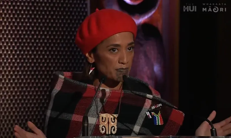 Oriini Kaipara speaks from behind a podium with her hands flayed out.