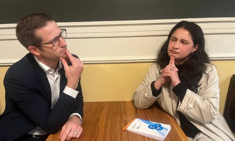 Chris Penk rubs his chin while the interviewer narrows her eyes and puts two fingers against her chin.
